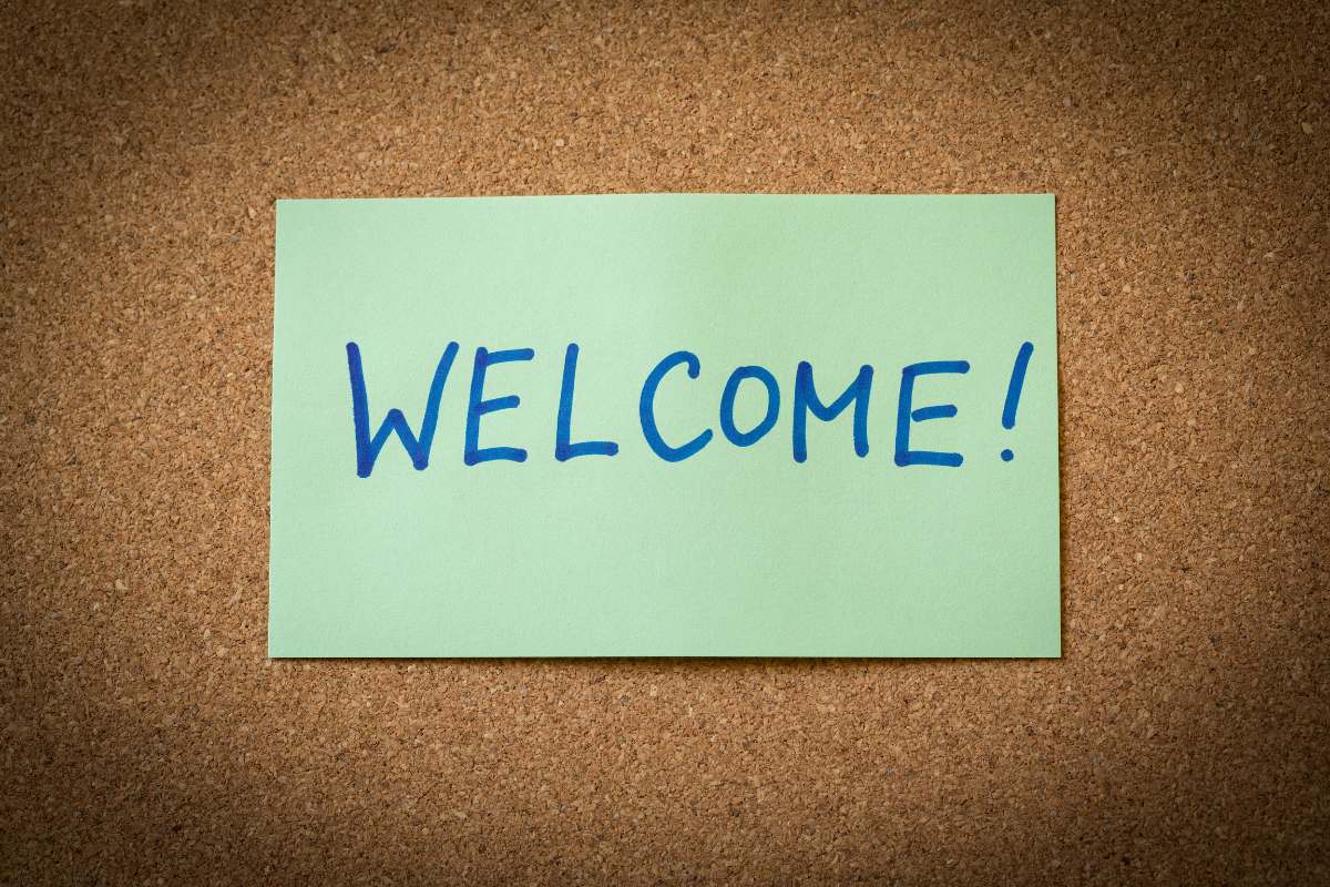 Welcome! written in in blue pen in capital letters on a light green post-it on a cork board