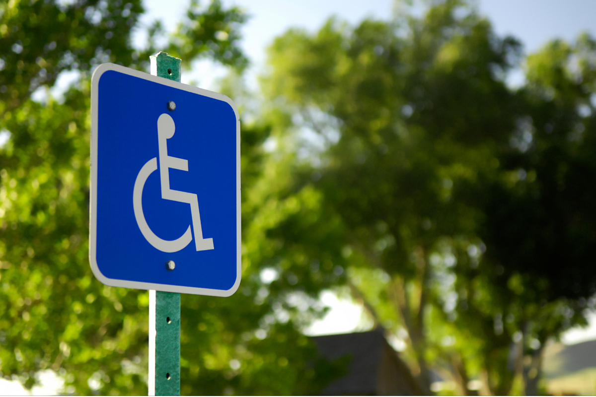 White on blue wheelchair symbol sign on a vertical post with green trees in the background