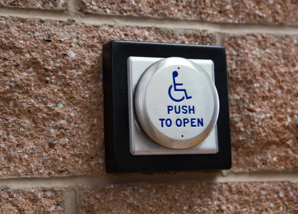 Round metal push button mounted on a brick wall, with blue wheelchair symbol and letters reading PUSH TO OPEN