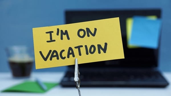 "I'M ON VACATION" printed in black sharpie ink on a yellow rectangular Post-It note in front of an out of focus laptop
