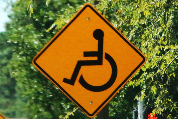Black wheelchair symbol on a diamond-shaped orange sign with leafy trees in background